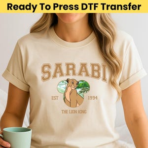 May include: Beige t-shirt with a brown graphic of Sarabi from The Lion King. The graphic includes the name "SARABI" in a brown collegiate font, the text "EST 1994", and a cartoon image of Sarabi. The text "THE LION KING" is below the graphic.