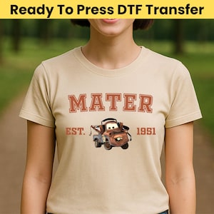May include: A tan t-shirt featuring a graphic of Mater from the movie Cars. The design includes the name "MATER" in a distressed font, the text "EST. 1951", and an image of the character. The shirt is short-sleeved.