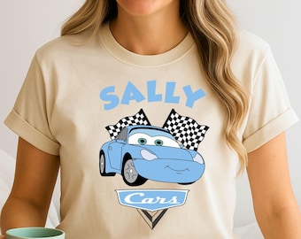 May include: Beige t-shirt featuring a light blue cartoon car with the name "SALLY" above it. The car has green eyes and is flanked by checkered flags. The word "Cars" is below the car. The text "Ready To Press DTF Transfer" is at the top.