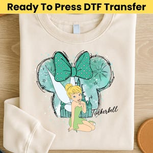 May include: Cream-colored sweatshirt with a DTF transfer featuring Tinkerbell, a green bow with white polka dots, and a castle silhouette. The text "Tinkerbell" is written in cursive. The words "Ready To Press DTF Transfer" are at the top.