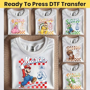 Puede incluir: Una colección de camisetas blancas con transferencias DTF Ready To Press de personajes de Nintendo como Mario, Peach, Bowser, Luigi, Donkey Kong y Yoshi. Cada camiseta muestra un personaje, su nombre en cursiva y un patrón de cuadros.