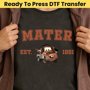 May include: Dark gray t-shirt featuring the word "MATER" in orange block letters above a cartoon image of Mater from the movie Cars. The text "EST. 1951" is below the image. The words "Ready To Press DTF Transfer" are at the top.