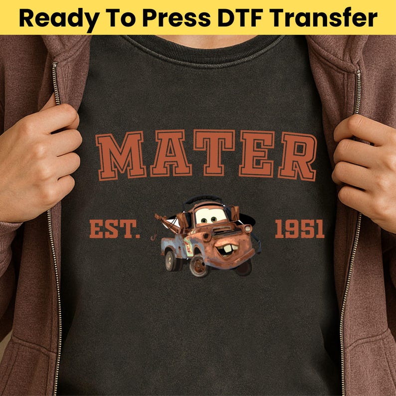 May include: Dark gray t-shirt featuring the word "MATER" in orange block letters above a cartoon image of Mater from the movie Cars. The text "EST. 1951" is below the image. The words "Ready To Press DTF Transfer" are at the top.