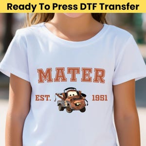 May include: White t-shirt featuring a graphic of Mater from the movie Cars. The design includes the name "MATER" in orange block letters, the text "EST. 1951", and an image of the character Mater.