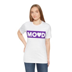 Könnte beinhalten: Ein weißes T-Shirt mit kurzen Ärmeln und einer rechteckigen violetten Grafik auf der Brust. Innerhalb des violetten Rechtecks ist das Wort MOOD in weißen Großbuchstaben gedruckt, wobei ein weißes Herz den Buchstaben O ersetzt.