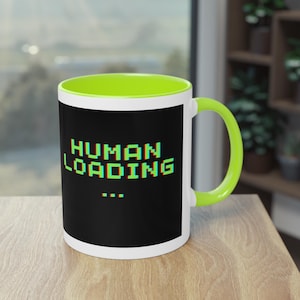 Könnte beinhalten: Ein weißer Keramikbecher mit hellgrünem Innenraum und Henkel. Die Außenseite zeigt eine schwarze rechteckige Grafik mit dem Text "HUMAN LOADING..." in einer verpixelten Glitch-Schriftart mit grünen und roten Farbverschiebungen.