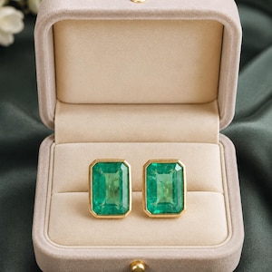 Puede incluir: Un par de pendientes rectangulares de esmeralda engastados en biseles de oro amarillo. Los pendientes se muestran dentro de un joyero de terciopelo beige claro abierto con un cierre de botón dorado, resaltando las piedras verdes.