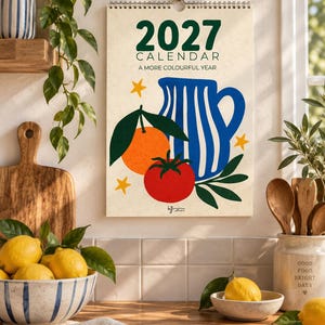 Könnte beinhalten: Ein Wandkalender für 2027 mit cremefarbenem Hintergrund, der einen blau-weiß gestreiften Krug, eine Orange, eine rote Tomate und gelbe Sterne zeigt. Der Text lautet 2027 CALENDAR A MORE COLOURFUL YEAR. Der Kalender hat eine Spiralbindung.