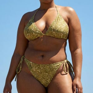 May include: A two-piece bikini set in a light olive green color with a subtle floral pattern. The top is a triangle style with adjustable straps, and the bottoms are a classic cut with tie sides. The background is a clear blue sky.