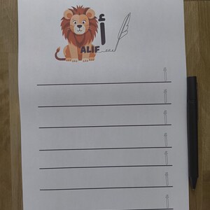 May include: White paper with black lines for writing practice. A cartoon lion illustration with the word "ALIF" in Arabic script is at the top. A black pen rests on the right side of the paper.