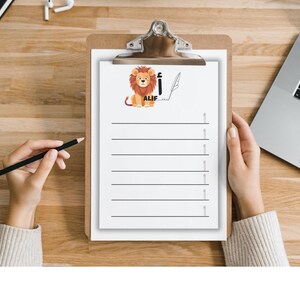 May include: A clipboard with a white paper featuring a cartoon lion illustration and the word "ALIF". The paper has horizontal lines for writing. A hand holds a black pencil, and another hand holds the clipboard.