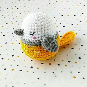 May include: A crocheted bird toy with a white head, grey wings, and a yellow body and tail. The bird has a small pink mouth and black eyes. The bird is on a white surface with black, yellow, and blue polka dots.