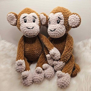 Könnte beinhalten: Zwei handgefertigte Häkelaffen. Die Affen sind braun mit hellbraunen Gesichtern, Händen und Füßen. Sie haben schwarz gestickte Augen und ein Lächeln. Die Affen sitzen auf einer weißen, flauschigen Oberfläche.