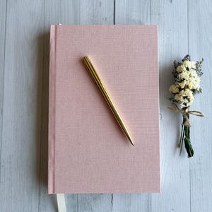 May include: A blush pink journal with a gold pen resting diagonally across the cover. A small bouquet of dried flowers with a twine tie sits to the right. The journal has a ribbon bookmark.