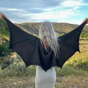 May include: A dark charcoal gray knit garment featuring large, pointed bat-like wing extensions that span outward from the shoulders and back, with visible ribbed detailing along the wing structure and a hood attached at the neckline.