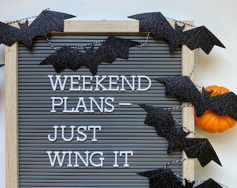 May include: A square gray felt letter board with a light wood frame, featuring white plastic letters that spell out WEEKEND PLANS - JUST WING IT. A string of black glittery bat cutouts is draped across the board, with a small orange pumpkin.