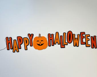 Puede incluir: Una guirnalda decorativa con el texto HAPPY HALLOWEEN en letras naranjas llamativas con bordes negros, unidas por un cordel fino. Una calabaza de fieltro naranja con una cara negra cosida reemplaza la letra O en la palabra HAPPY.