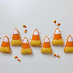 May include: A garland featuring seven felt candy corn shapes strung together on a thin white cord. Each candy corn piece consists of a white top, an orange middle section, and a yellow base, with visible stitching along the edges of each segment.