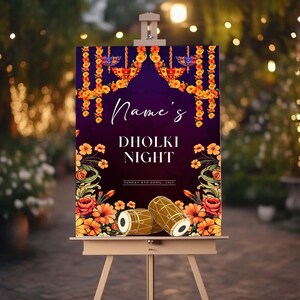 Peut inclure: Panneau d&eacute;coratif sur chevalet avec l'inscription "Name's Dholki Night" en &eacute;criture blanche. Le panneau pr&eacute;sente des illustrations florales et de tambours avec un fond violet fonc&eacute; et des accents dor&eacute;s. La date est imprim&eacute;e en bas.