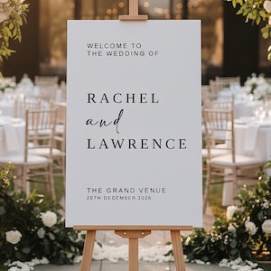 May include: A white wedding sign on a wooden easel, with the text "Welcome to the Wedding of Rachel and Lawrence". The venue and date are also displayed. The background features floral decorations and a reception setting.