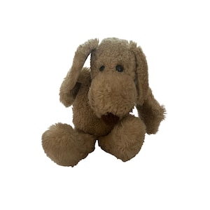 May include: A plush dog toy made of light brown, textured, fuzzy fabric. The stuffed animal features long, floppy ears, a rounded snout, and two small, dark, round eyes. It is depicted in a seated position with its front paws resting forward.