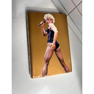 May include: A book with a gold cover featuring a color image of a blonde woman in a black bodysuit and fishnet stockings, holding a microphone. The woman is singing on a stage with a gold background.