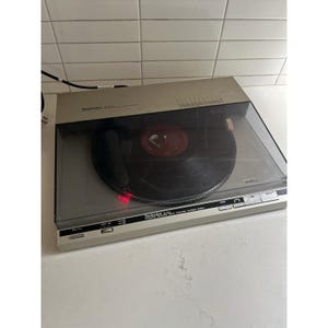 May include: A silver Technics SL-B2 frequency generator servo automatic turntable system with a black vinyl record on the platter. The device features a transparent dust cover, a pitch adjustment slider, and a red light indicator on the tonearm.
