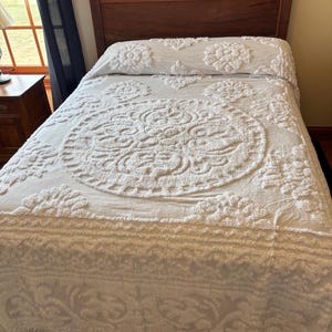 May include: A white bedspread with a raised, tufted floral and circular medallion pattern. The textile features a textured, chenille-like surface with a decorative fringe along the bottom edge, covering a bed with a dark wooden headboard.