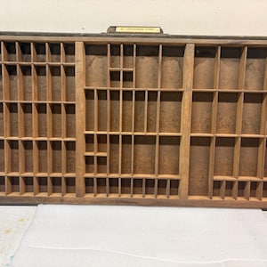 May include: A rectangular wooden printer's type case featuring numerous small, varied compartments for organizing metal letters. The frame is constructed from light brown wood and includes a small label at the top center that reads Cheltenham Gold.