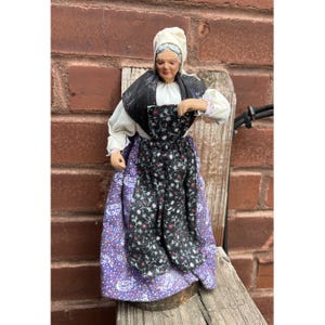 May include: A detailed figurine of an elderly woman in traditional attire. She wears a white head covering, a black shawl, and a black floral apron over a purple skirt. The figure is posed on a wooden surface against a brick background.