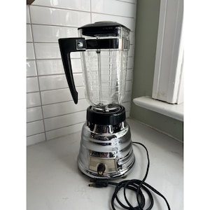 May include: A vintage chrome and glass blender with a black handle and lid. The clear glass pitcher has measurement markings. The blender sits on a white countertop with a black power cord.