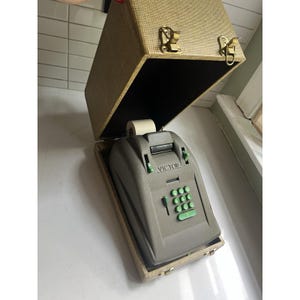May include: A vintage Victor adding machine with a grey plastic casing and bright green circular keys, housed in a textured beige carrying case with gold-toned metal latches. The device features a paper roll holder and a numeric keypad.