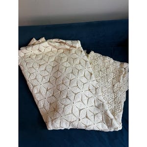 May include: Cream-colored, open-weave textile with a floral pattern. The fabric is folded, revealing the intricate design and texture. The textile is likely a decorative item, such as a tablecloth or throw.