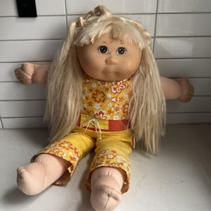 May include: A vintage Cabbage Patch Kid doll with long blonde hair and a soft, round face. The doll is wearing a yellow floral print outfit with an orange belt. The doll's arms are outstretched, and it is sitting down.