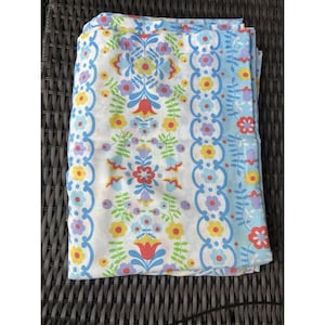 May include: Folded white fabric with a colorful floral pattern. The design features red, yellow, blue, and purple flowers, with green leaves and stems. The edges have a blue border with a repeating floral motif.