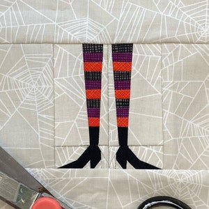 Puede incluir: Primer plano de un cuadrado de tela con un patrón de telaraña. Un cuadrado de tela más pequeño con un diseño de piernas de bruja está cosido en el cuadrado más grande. Hay tres clips de tela y un par de tijeras sobre una superficie blanca. El texto "HAPPYSEWLUCKY" está impreso en la tela.