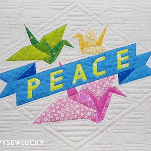 May include: A white quilt with a blue banner that says "PEACE" in yellow letters. The banner is surrounded by four origami paper cranes in green, yellow, and pink.