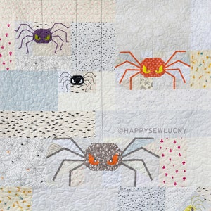 Könnte beinhalten: Eine weiße Steppdecke mit Patchwork-Muster, die vier Spinnen mit unterschiedlich farbigen Körpern und Beinen zeigt. Die Steppdecke hat einen rosa-weißen Rand. Der Text "@happyssewlucky" befindet sich in der rechten unteren Ecke der Steppdecke.