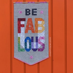 May include: A grey fabric banner with the words "BE FABULOUS" in rainbow colours. The banner is hanging on a bright orange wall.