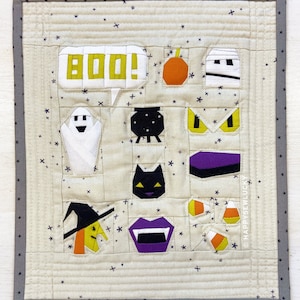 May include: A beige fabric wall hanging with a grey border. The wall hanging features a variety of Halloween-themed images, including a ghost, a witch, a vampire, a black cat, a cauldron, a pumpkin, candy corn, and a coffin. The text "BOO!" is in a yellow speech bubble.
