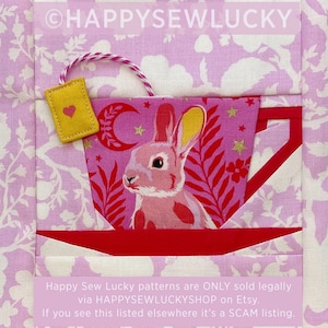 May include: A fabric quilt block featuring a central teacup graphic with a pink rabbit illustration, a yellow tea bag with a heart, and a red saucer. The background is light purple with white floral patterns. Text reads HAPPYSEWLUCKY.