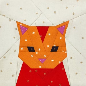 May include: A geometric orange cat face quilt block with black eyes, pink inner ears, and a red chest. The background is a light beige fabric with small white snowflakes. The text "HAPPYSEWLUCKY" is visible.