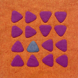 May include: A grid of purple felt triangles on an orange fabric background. One triangle in the center is a different color, a dark blue.