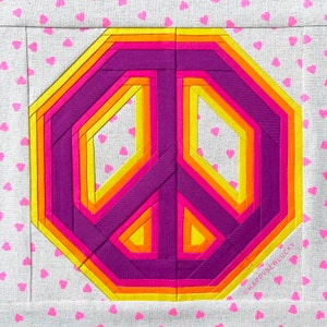 May include: A colorful peace sign made of fabric strips in shades of yellow, orange, pink, and purple. The peace sign is on a white background with a pattern of pink hearts.