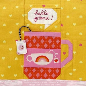 May include: A yellow fabric quilt block featuring a central pink and orange patterned mug with a rainbow graphic. A small white tea bag with a pink smiley face hangs from a black and white string. The text hello friend is embroidered above.