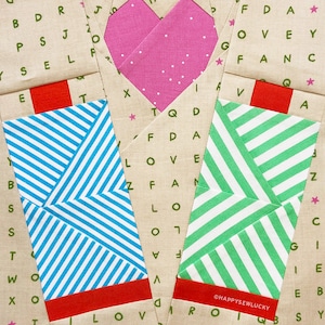 Könnte beinhalten: Drei Stoffquadrate mit einem rot-grün-blau gestreiften Muster. Jedes Quadrat hat eine rote Klappe oben und eine weiße Klappe unten. Die Quadrate sind in Dreiecksform angeordnet, mit einem rosa Herz in der Mitte. Das Herz hat weiße Punkte. Die Stoffquadrate haben Buchstaben darauf gedruckt, darunter die Wörter "LOVE" und "HAPPYSEWLUCKY".