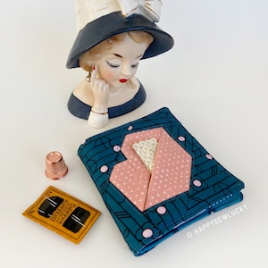 Può includere: Una pochette in tessuto blu turchese con un design a forma di cuore rosa e pois bianchi. La pochette è chiusa e ha un'etichetta bianca con il testo "HAPPYSEWLUCKY".
