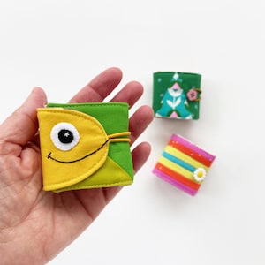 Pode incluir: Três pequenos cadernos cobertos de tecido. Um é amarelo com uma aba verde e um olho preto com uma pupila branca. Outro é verde com uma flor rosa e um desenho branco. O último é rosa com listras arco-íris e uma flor branca.