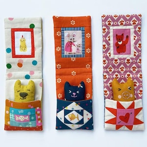 Może przedstawiać: Trzy ręcznie robione zakładki do książek z fantazyjnymi wzorami. Każda zakładka posiada małą kieszonkę, w której znajduje się pluszowe zwierzątko. Zakładki mają różne wzory i kolory, w tym kropki, nadruki kwiatowe i wzory patchworkowe. Każda zakładka ma oprawioną ilustrację.