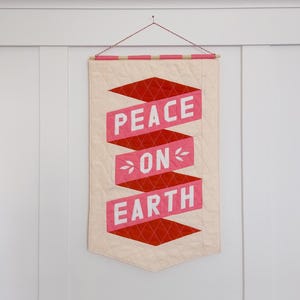 May include: A white fabric banner with a red and pink quilted design. The banner has the words "Peace on Earth" written in white on the red and pink fabric.